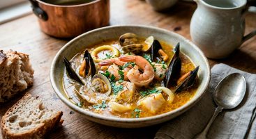 Un primer plano de un cuenco de cerámica rústico y artesanal lleno de sopa de mariscos dorada. Mariscos abiertos como mejillones, almejas, gambas y cubos de pescado irregulares flotan en el caldo azafranado. Perejil fresco picado está espolvoreado por encima. El cuenco está sobre una mesa de madera rústica.