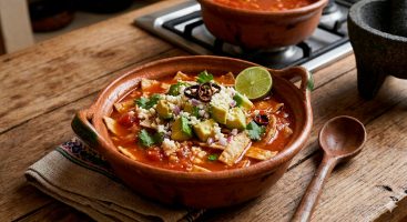 Un tazón de barro lleno de sopa de tortilla con caldo rojo, coronado con abundantes cubos de aguacate, queso fresco, anillos de chile seco, cilantro y una rodaja de limón, sobre un paño de tela y una mesa rústica de madera.