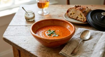 Un tazón de cerámica rústica lleno de una sopa de tomate rostizado espesa y de color rojo intenso, sobre una mesa de madera oscura. La sopa está decorada artísticamente con tres hojas enteras de albahaca verde fresca en el centro. Al lado, una cuchara de plata vintage descansa sobre una servilleta de lino deshilachada. De fondo, desenfocados, se ven pan sourdough artesanal y una copa de vino.