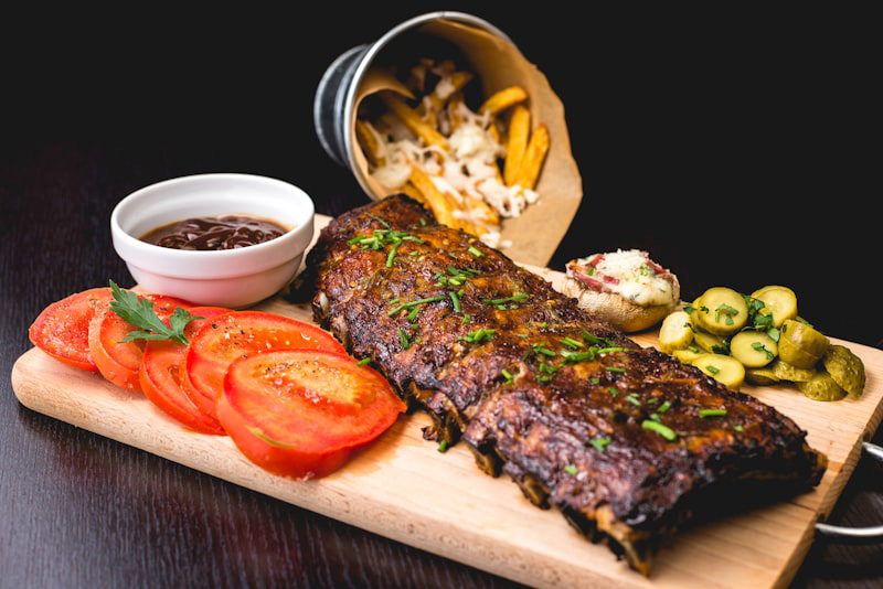 Un plano cenital en ángulo medio de un costillar de cerdo asado y glaseado con barbacoa, espolvoreado con cebollino picado, servido sobre una tabla de cortar de madera clara con mango de metal. A la izquierda, hay cuatro rodajas de tomate fresco sazonadas con sal y pimienta y una ramita de perejil. Un cuenco de cerámica blanca lleno de salsa barbacoa oscura y espesa con un cuchillo de untar descansa arriba a la izquierda. A la derecha, una guarnición de pepinillos encurtidos en rodajas con perejil picado. Al fondo, un cubo metálico volcado sobre papel de estraza, del que salen patatas fritas cubiertas de queso derretido. El fondo es una superficie de madera oscura y borrosa con una atmósfera de pub rústico. La iluminación es natural y uniforme.